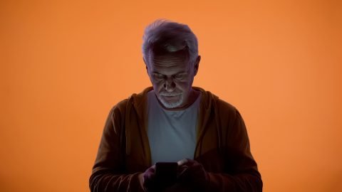 Upset Old Man Typing Message On Smartphone, Orange Background, Vision Problem