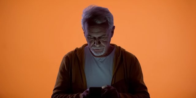 Upset old man typing message on smartphone, orange background, vision problem